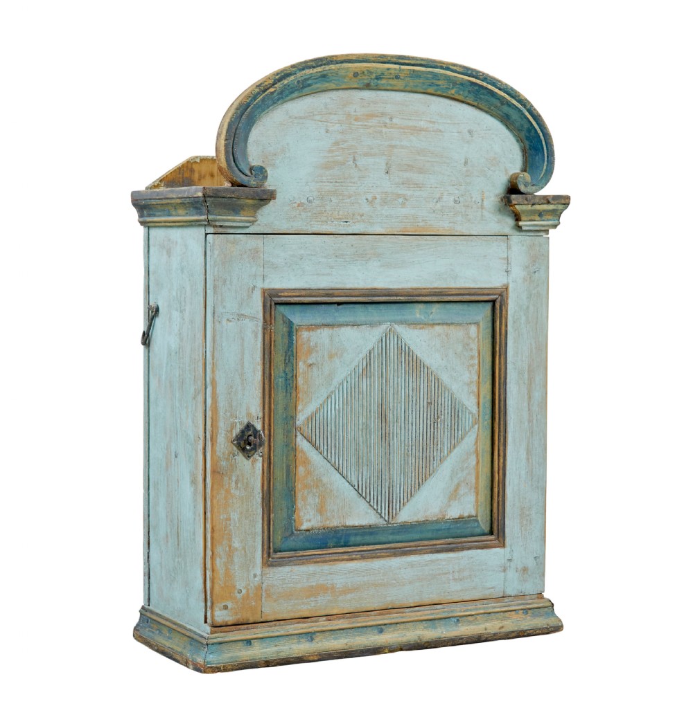 A 19th-century Finnish painted pine wall cupboard featuring weathered light blue paint, an arched pediment crest, and a door panel carved with a diamond pattern.