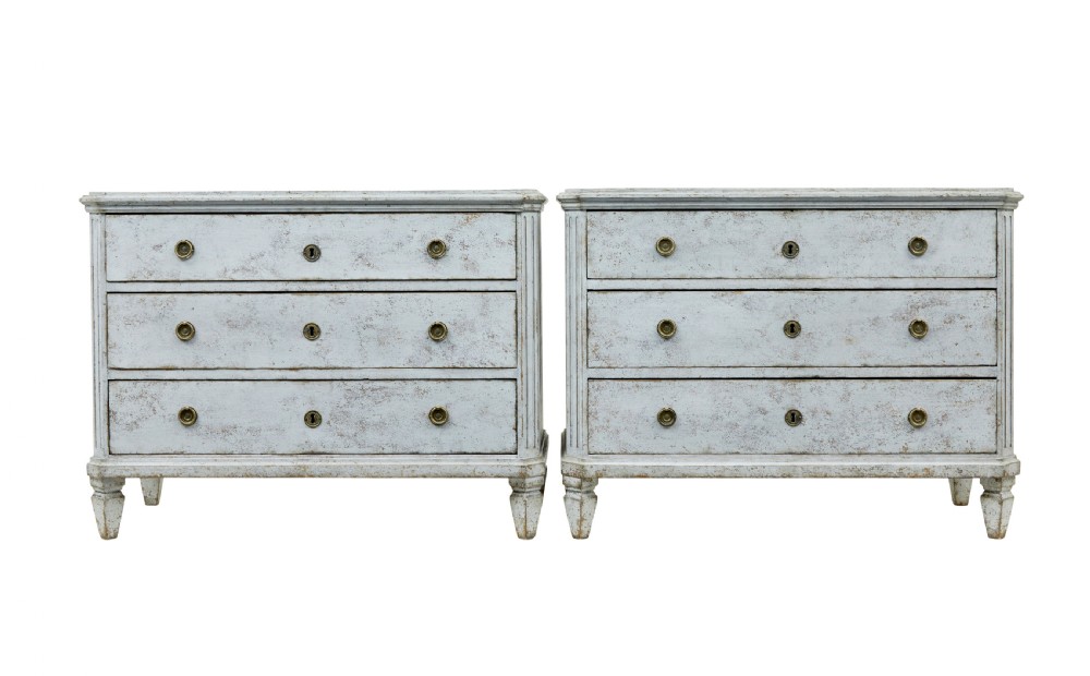 A pair of 19th-century Gustavian style painted chests of drawers with three drawers each, brass pulls, fluted side columns, and distressed pale grey paint.