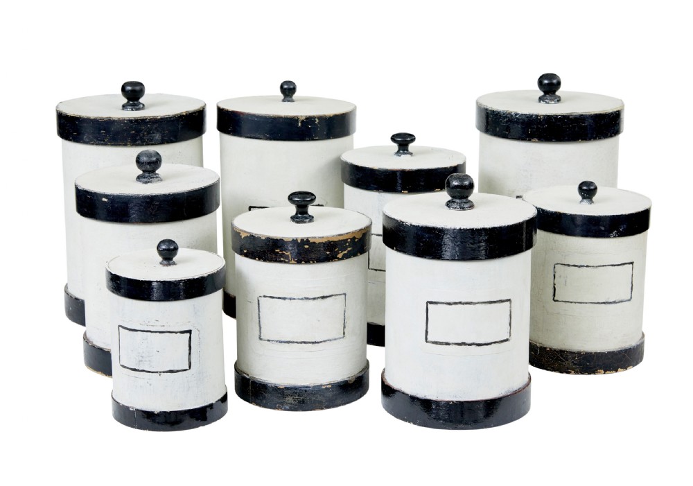 A set of eight cylindrical off-white painted wooden jars with black bases, black rims, turned black lid knobs, and painted rectangular label borders.