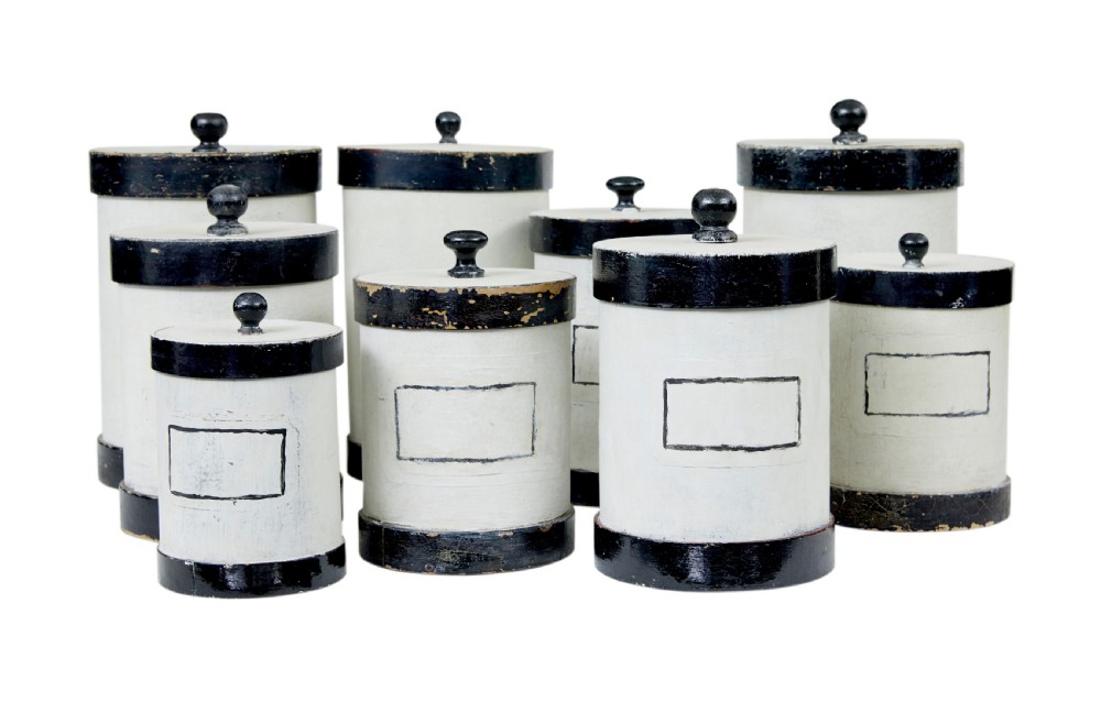 A set of eight cylindrical off-white painted wooden jars with black bases, black rims, turned black lid knobs, and painted rectangular label borders. View II