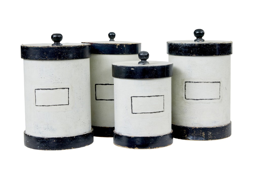 A set of eight cylindrical off-white painted wooden jars with black bases, black rims, turned black lid knobs, and painted rectangular label borders. View III