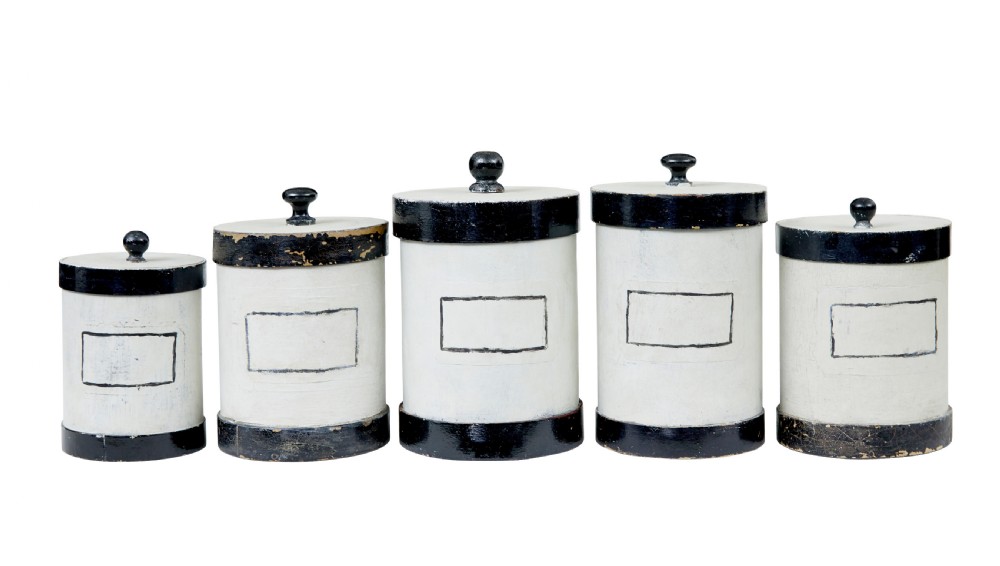 A set of eight cylindrical off-white painted wooden jars with black bases, black rims, turned black lid knobs, and painted rectangular label borders. View IV