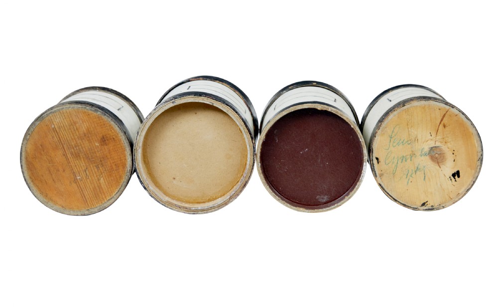 A set of eight cylindrical off-white painted wooden jars with black bases, black rims, turned black lid knobs, and painted rectangular label borders. View V