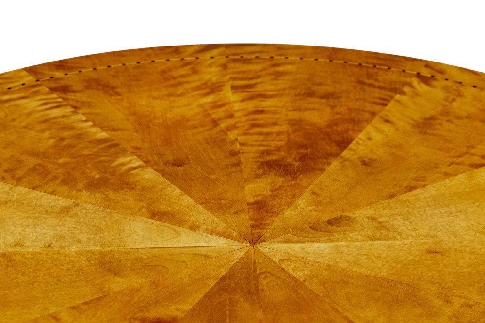 A circular golden flame birch centre table featuring a starburst-patterned veneer top, decorative inlaid rim border, sweeping concave pedestal stem, and quadform base with bun feet. View VI