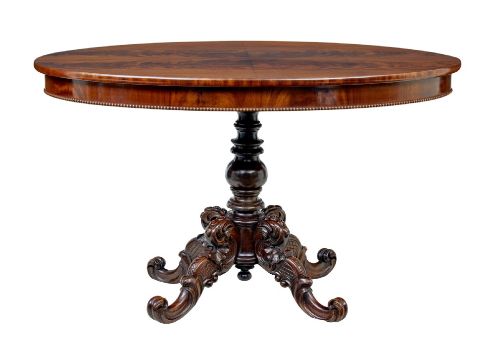 A 19th-century oval wood centre table featuring a flame mahogany veneered top, beaded apron moulding, turned central column, and carved scroll feet.