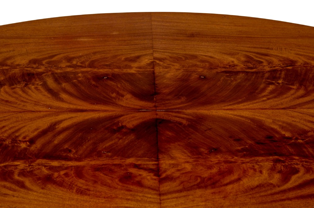 A 19th-century oval wood centre table featuring a flame mahogany veneered top, beaded apron moulding, turned central column, and carved scroll feet. View VII