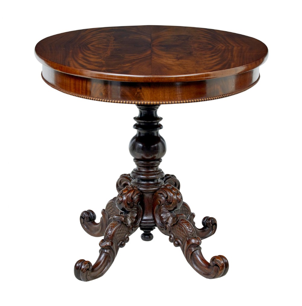 A 19th-century oval wood centre table featuring a flame mahogany veneered top, beaded apron moulding, turned central column, and carved scroll feet. View IX