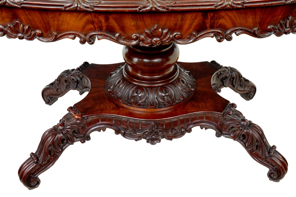 An antique 19th-century oval mahogany centre table featuring a carved apron, a central turned pedestal stem, and a carved four-legged base with scroll feet. View V
