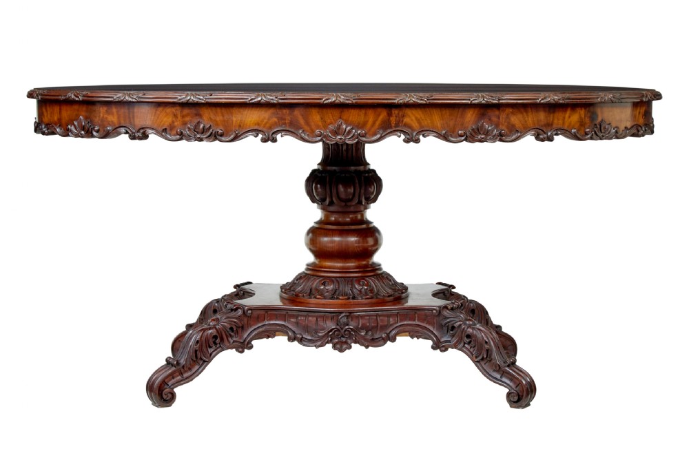An antique 19th-century oval mahogany centre table featuring a carved apron, a central turned pedestal stem, and a carved four-legged base with scroll feet. View IX