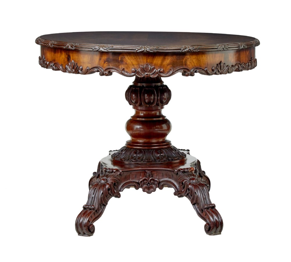An antique 19th-century oval mahogany centre table featuring a carved apron, a central turned pedestal stem, and a carved four-legged base with scroll feet. View X