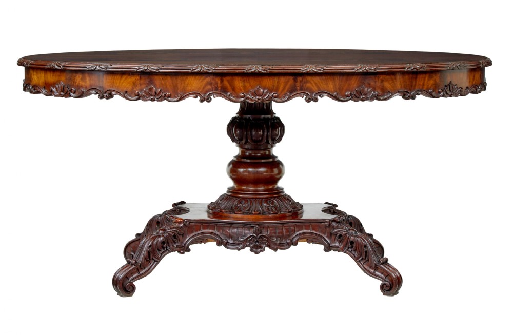 An antique 19th-century oval mahogany centre table featuring a carved apron, a central turned pedestal stem, and a carved four-legged base with scroll feet. View XI