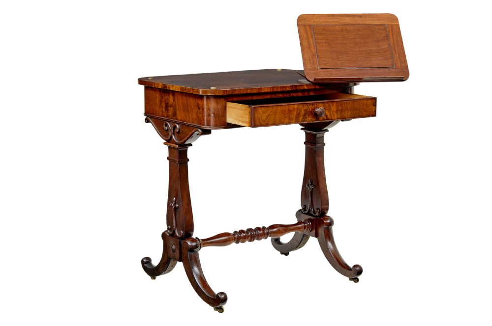 A mid-19th century dark mahogany writing table featuring an adjustable reading slope on top, a single open frieze drawer, scrolled side supports, turned stretcher, and brass castors.