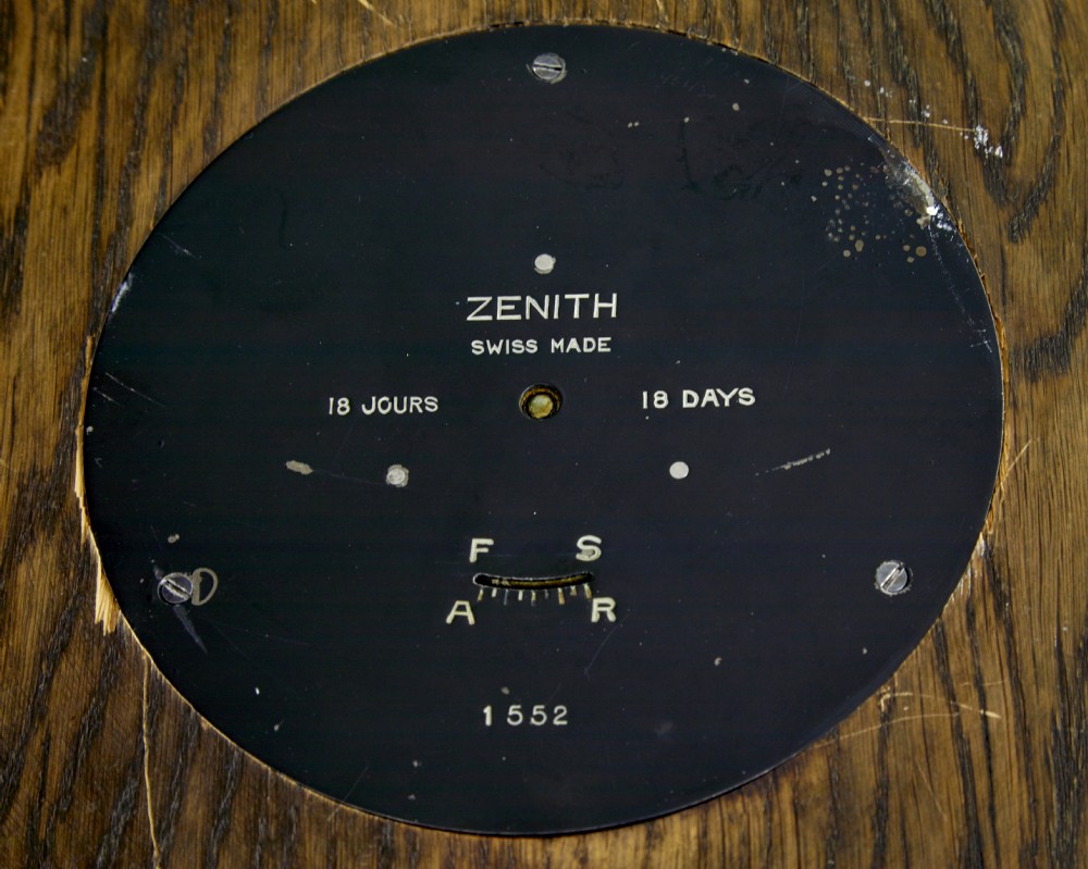 A circular Art Deco wall clock crafted from medium brown oak featuring applied brass Arabic numerals and metallic hands. View VI