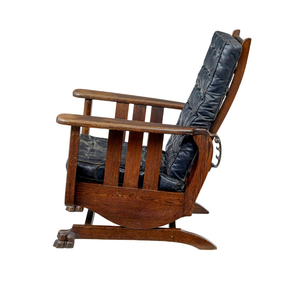 An early 20th-century dark oak platform rocking chair featuring button-tufted black leather cushions and carved front feet. View III