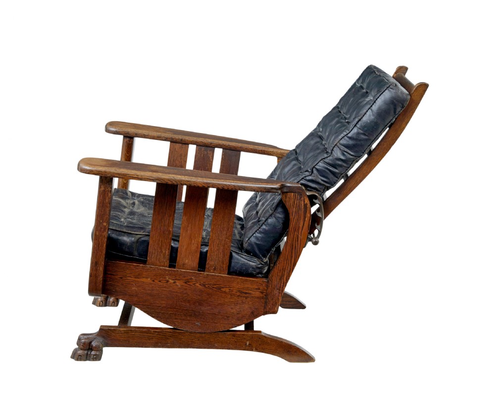 An early 20th-century dark oak platform rocking chair featuring button-tufted black leather cushions and carved front feet. View IV