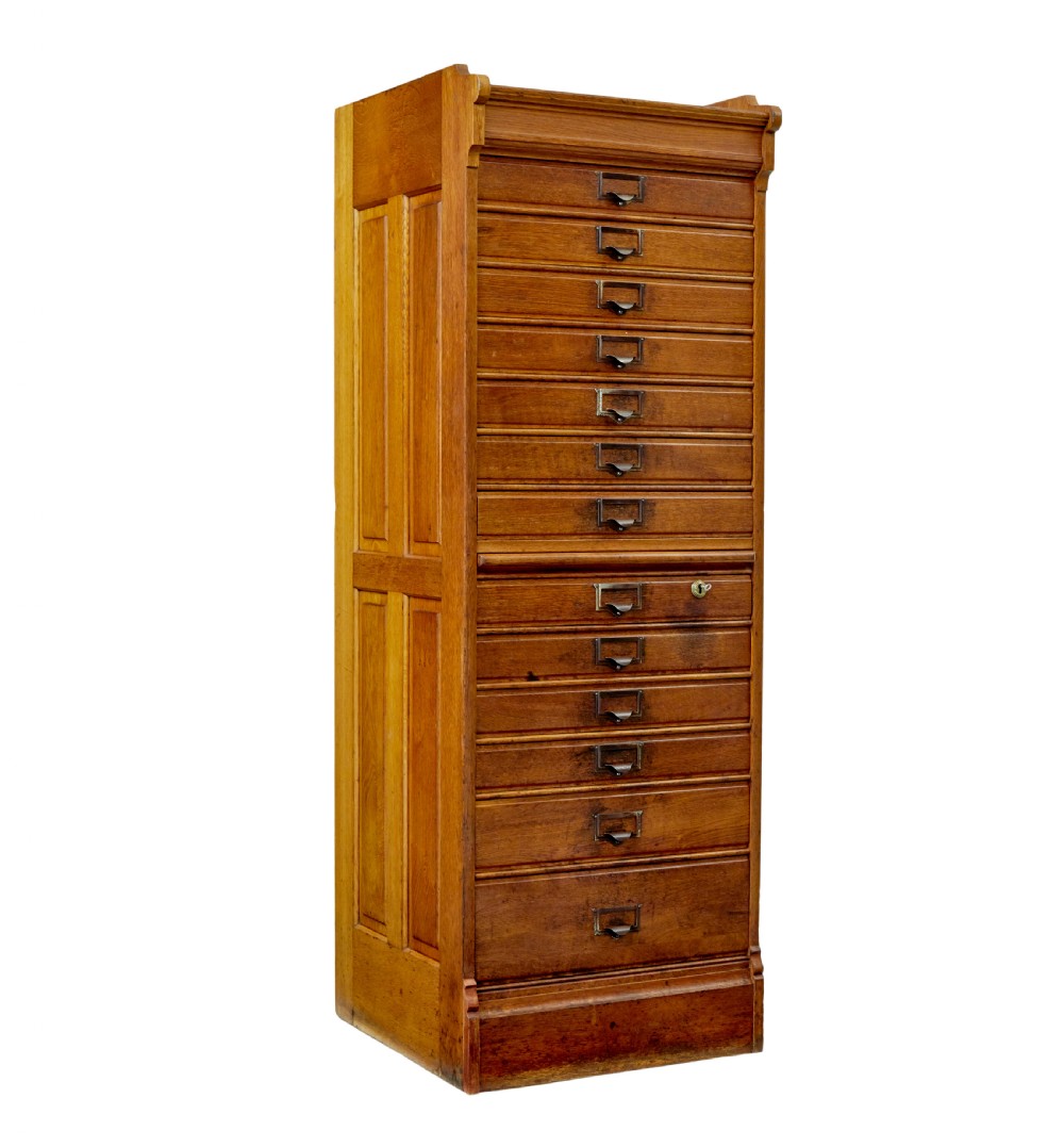 A tall early 20th-century solid oak filing chest featuring fourteen drawers with brass cup handles and label frames, flanked by paneled sides.