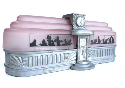 fig 1. An Art Deco style lamp featuring a stepped pink frosted glass body decorated with black silhouetted figures dining inside windows, framed by a silver-coloured cast metal base and central architectural doorway with a clock motif.