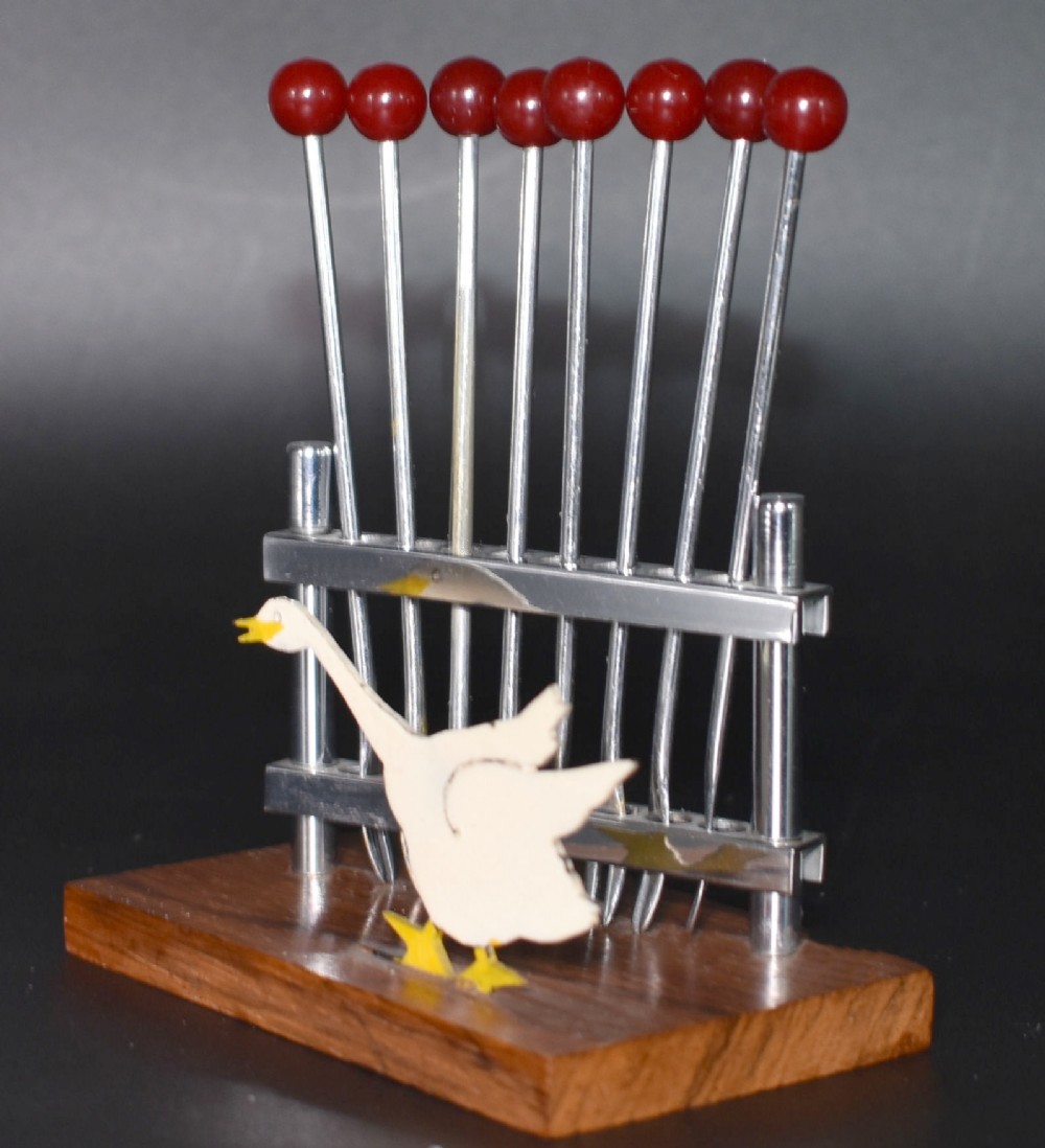 An Art Deco cocktail pick set featuring red Bakelite-topped chrome sticks resting in a wooden stand with a white goose detail.