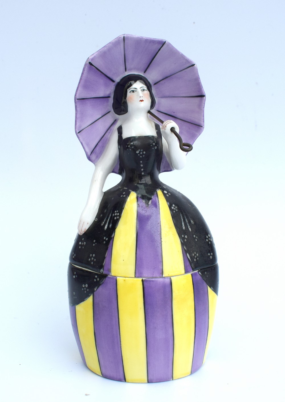 An Art Deco ceramic bonbonniere shaped as a woman holding a purple parasol and wearing a black corset bodice with a purple and yellow striped dress.