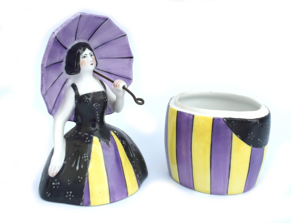 An Art Deco ceramic bonbonniere shaped as a woman holding a purple parasol and wearing a black corset bodice with a purple and yellow striped dress. View II