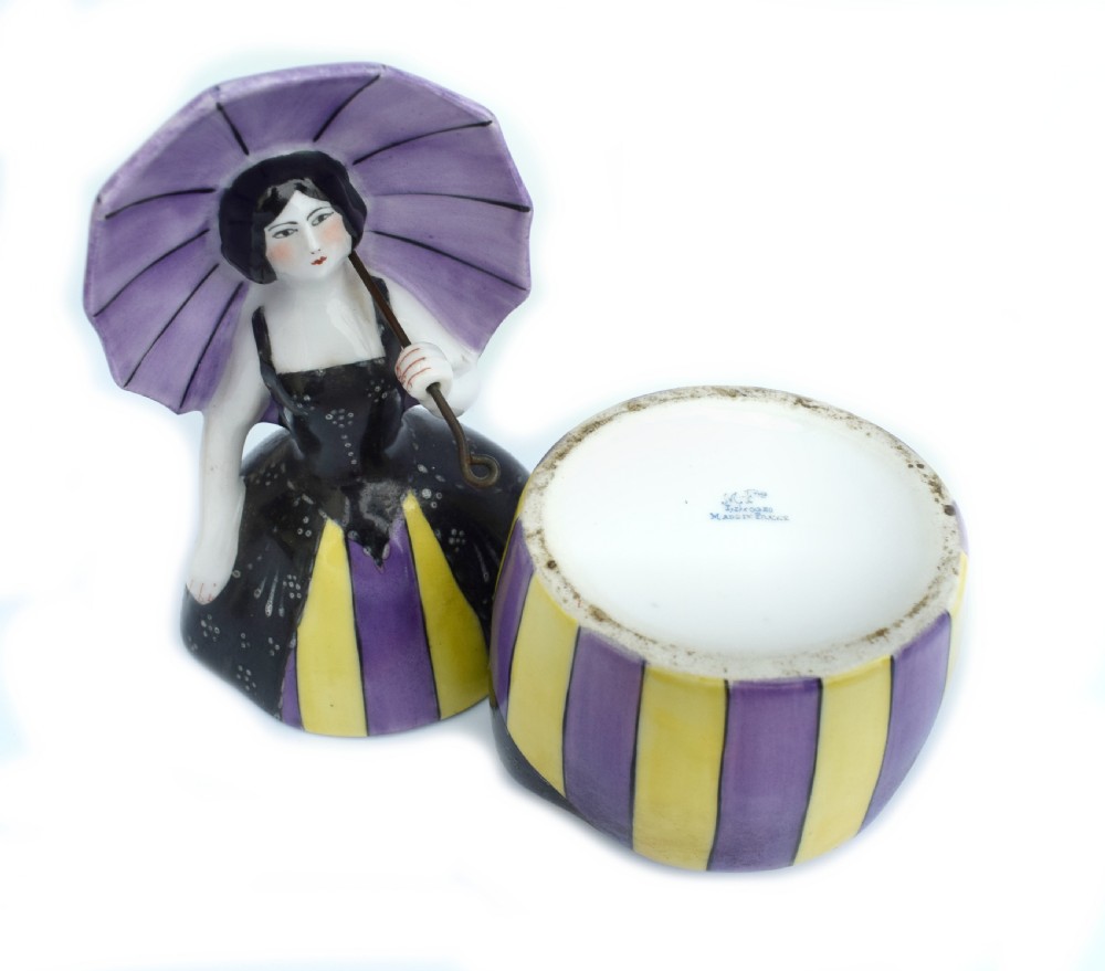 An Art Deco ceramic bonbonniere shaped as a woman holding a purple parasol and wearing a black corset bodice with a purple and yellow striped dress. View III