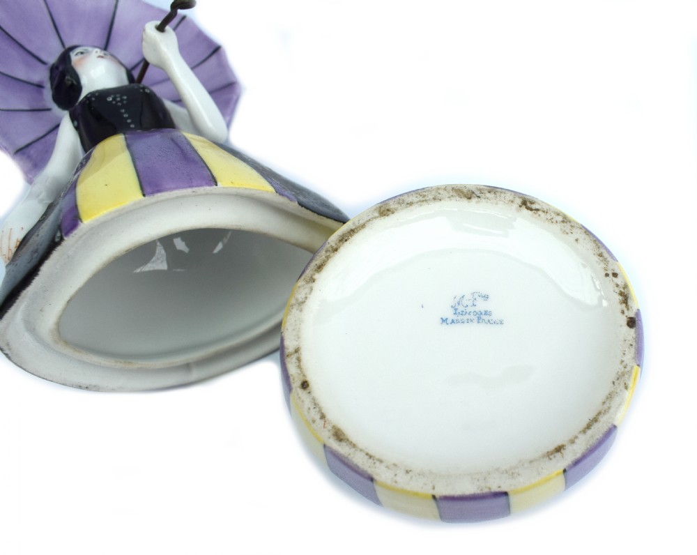 An Art Deco ceramic bonbonniere shaped as a woman holding a purple parasol and wearing a black corset bodice with a purple and yellow striped dress. View VI