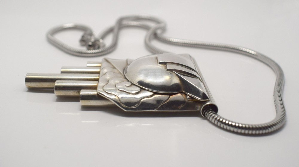 An Art Deco style silver-coloured chrome pendant featuring geometric stepped tubes and a central semicircle motif attached to a snake chain. View VII