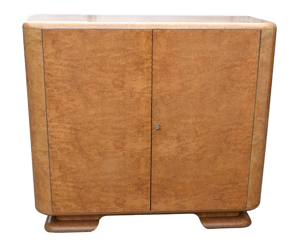 A two-door 1930s Art Deco bedroom cabinet featuring curved corners, figured blonde wood veneer, and low plinth feet.