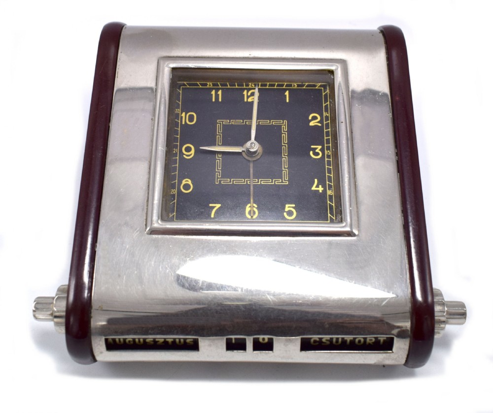 An Art Deco Hungarian desk clock featuring burgundy Bakelite side panels, a polished chrome casing, a square black dial, and integrated day and month calendar displays. View V