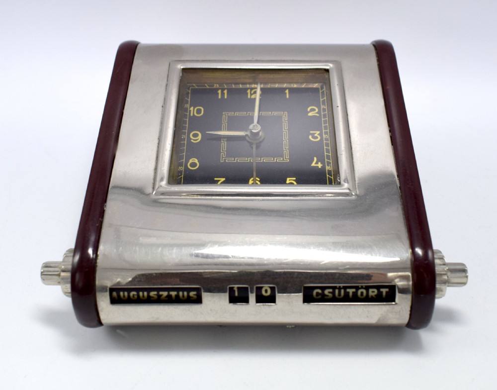 An Art Deco Hungarian desk clock featuring burgundy Bakelite side panels, a polished chrome casing, a square black dial, and integrated day and month calendar displays. View VI