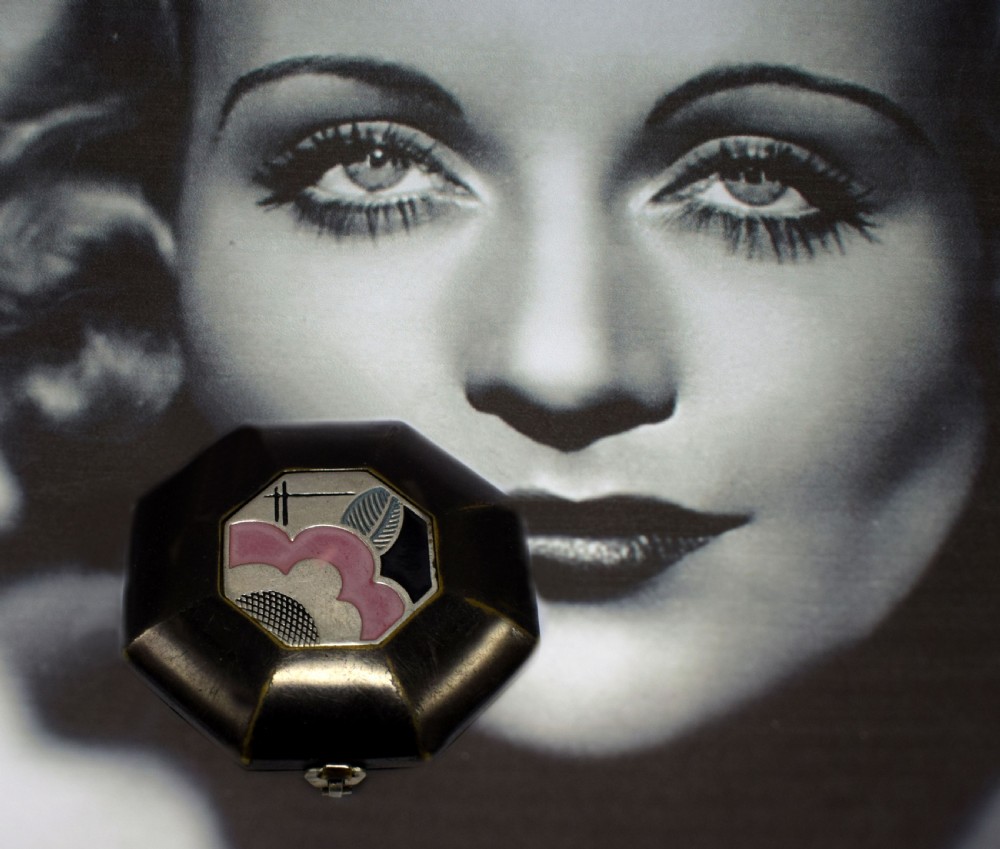 An octagonal Art Deco powder compact featuring a dark casing with a central enamelled plaque displaying pink, black, and silver geometric motifs. View IV