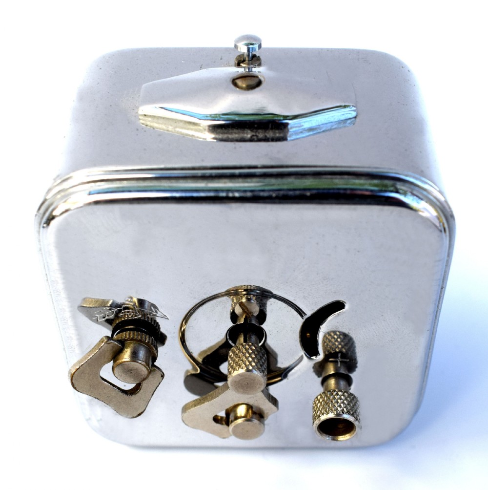 A square French Art Deco chrome alarm clock with stylized yellow numerals, a silver dial, and a top shut-off button. View III