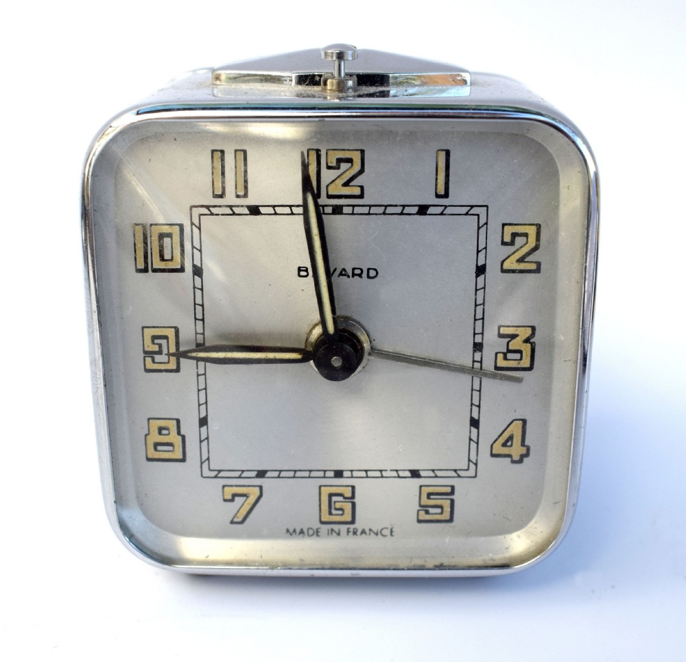 A square French Art Deco chrome alarm clock with stylized yellow numerals, a silver dial, and a top shut-off button. View IV