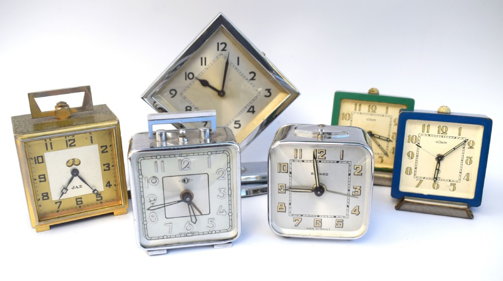 A square French Art Deco chrome alarm clock with stylized yellow numerals, a silver dial, and a top shut-off button. View VII