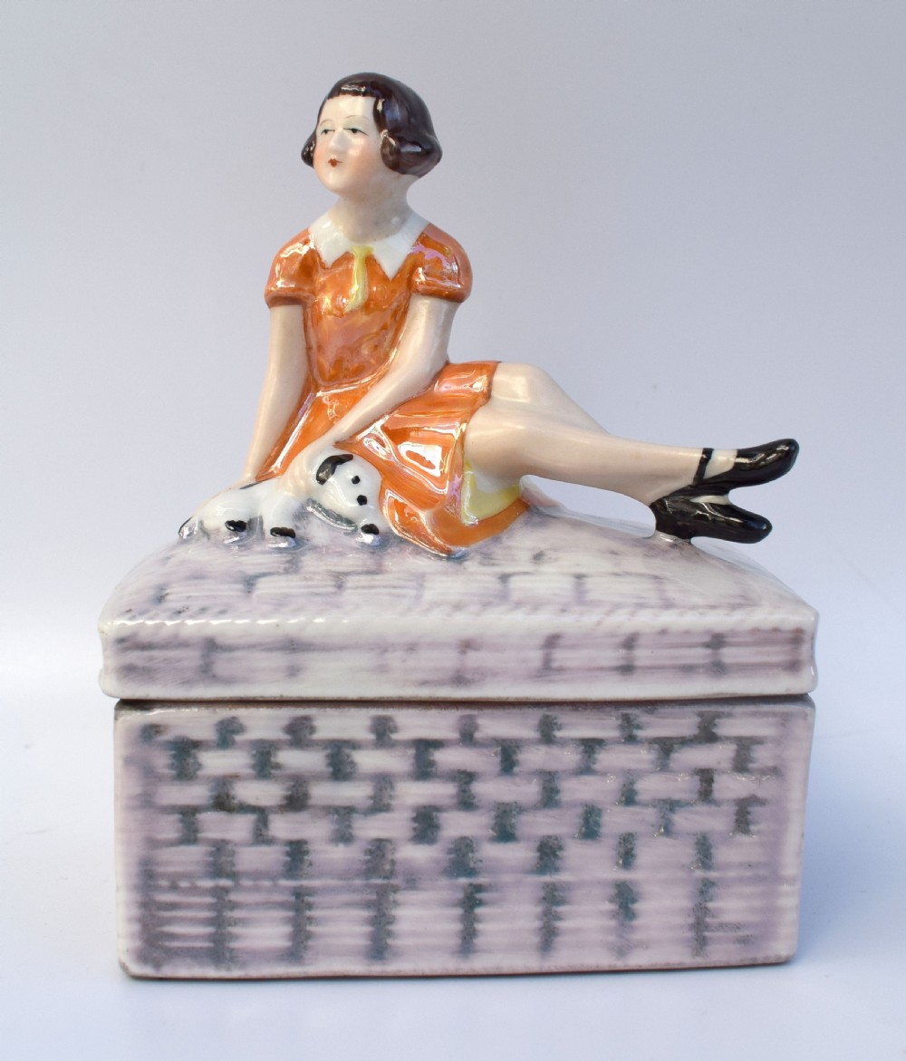 A glazed ceramic Art Deco trinket box featuring a lid adorned with a figurine of a seated girl in an orange dress with a puppy, over a basketweave patterned base.