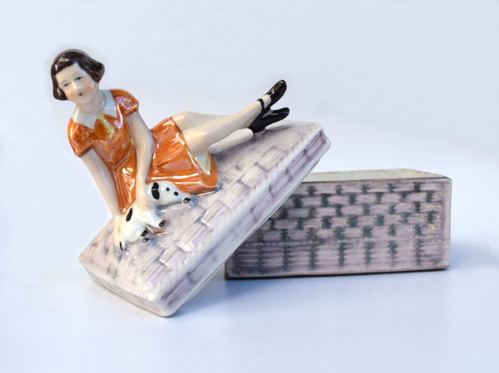 A glazed ceramic Art Deco trinket box featuring a lid adorned with a figurine of a seated girl in an orange dress with a puppy, over a basketweave patterned base. View II