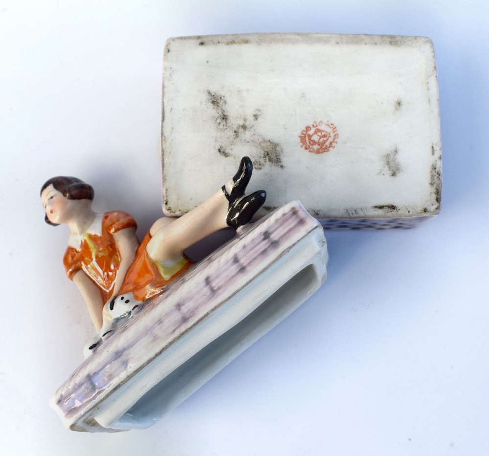 A glazed ceramic Art Deco trinket box featuring a lid adorned with a figurine of a seated girl in an orange dress with a puppy, over a basketweave patterned base. View III