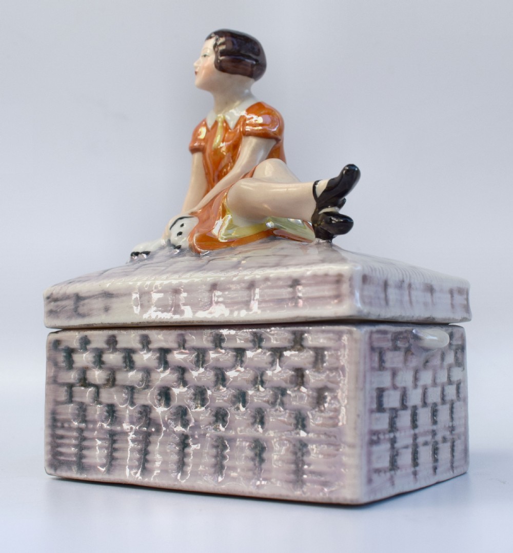 A glazed ceramic Art Deco trinket box featuring a lid adorned with a figurine of a seated girl in an orange dress with a puppy, over a basketweave patterned base. View IV