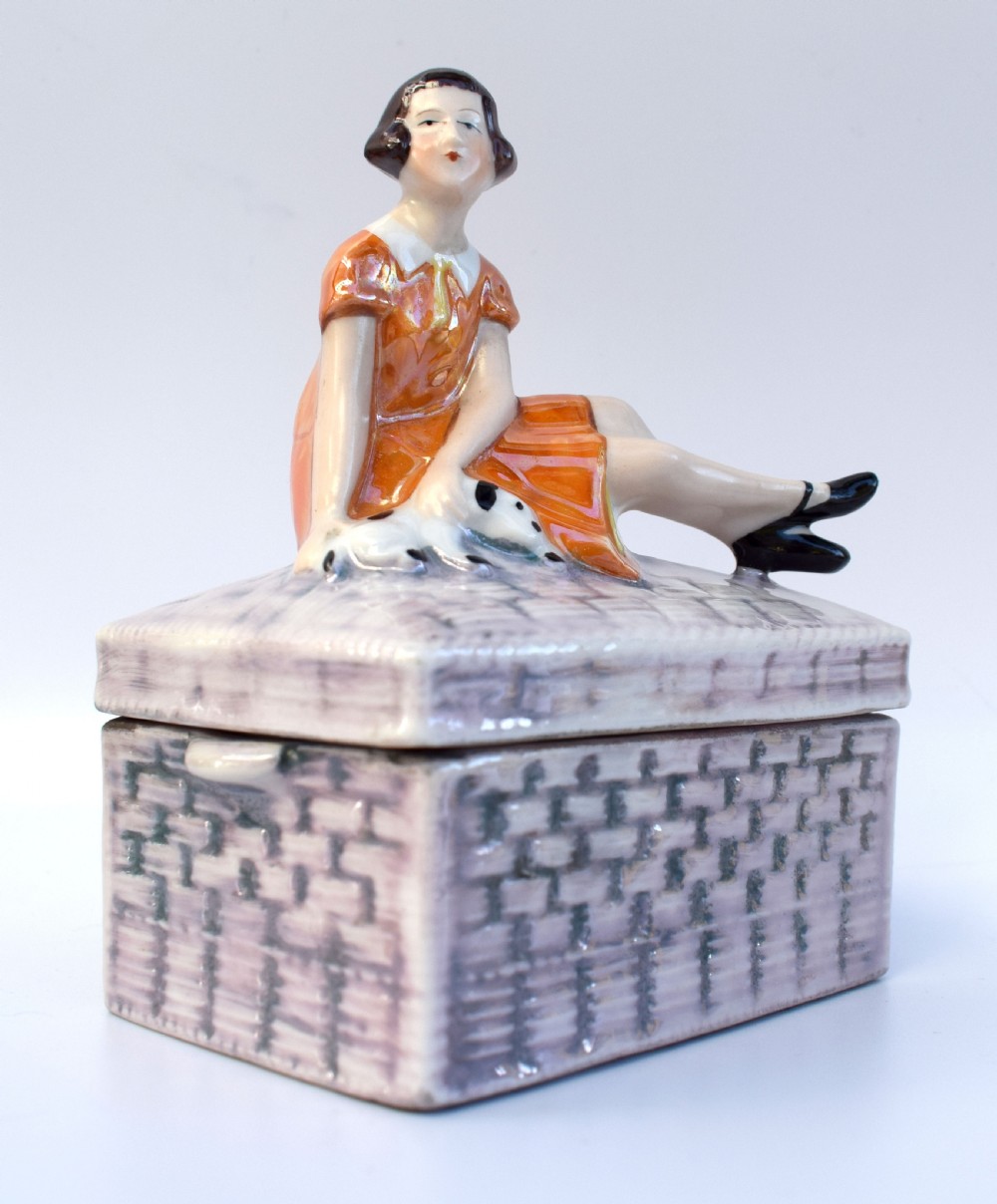 A glazed ceramic Art Deco trinket box featuring a lid adorned with a figurine of a seated girl in an orange dress with a puppy, over a basketweave patterned base. View V