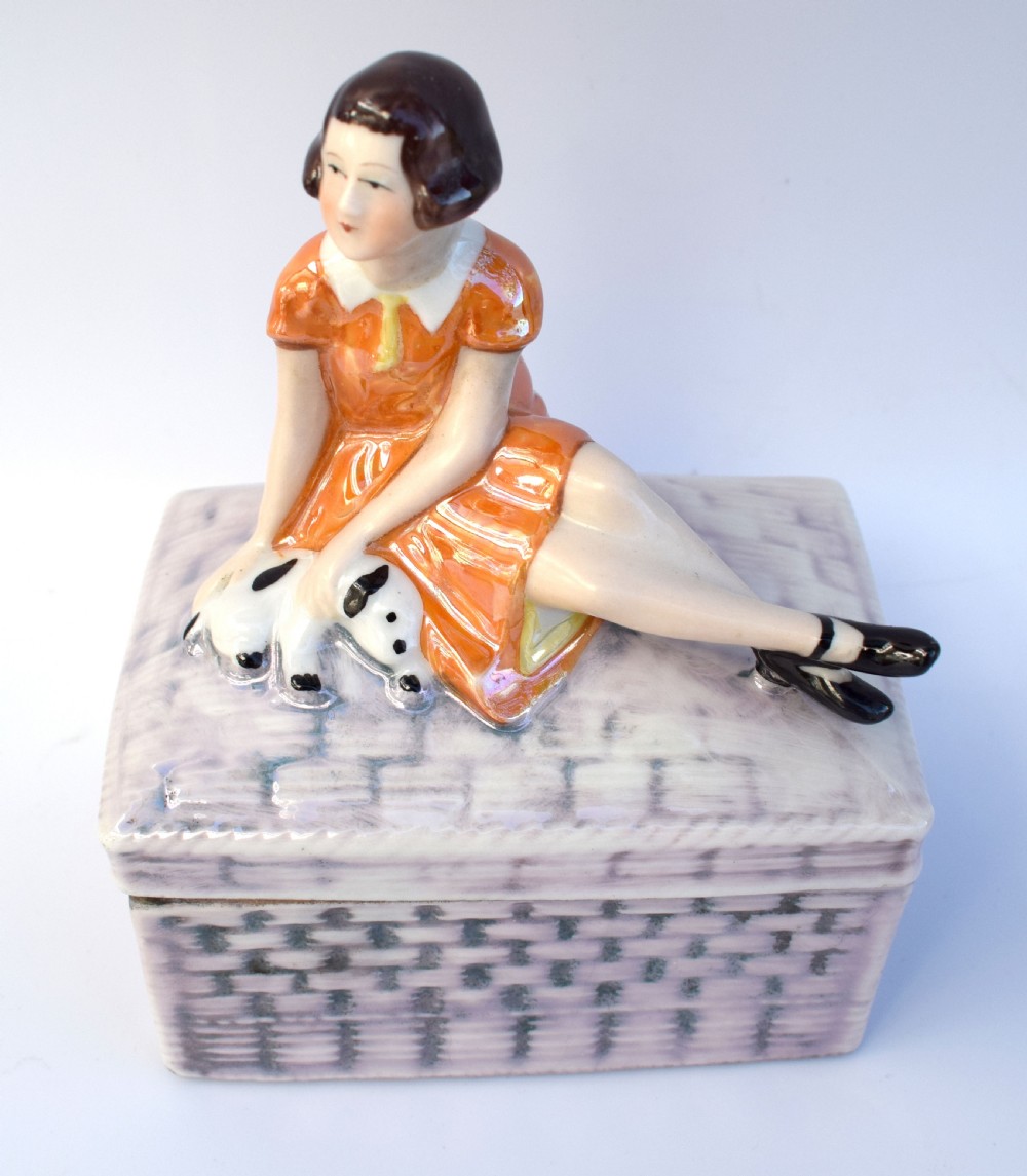 A glazed ceramic Art Deco trinket box featuring a lid adorned with a figurine of a seated girl in an orange dress with a puppy, over a basketweave patterned base. View VI