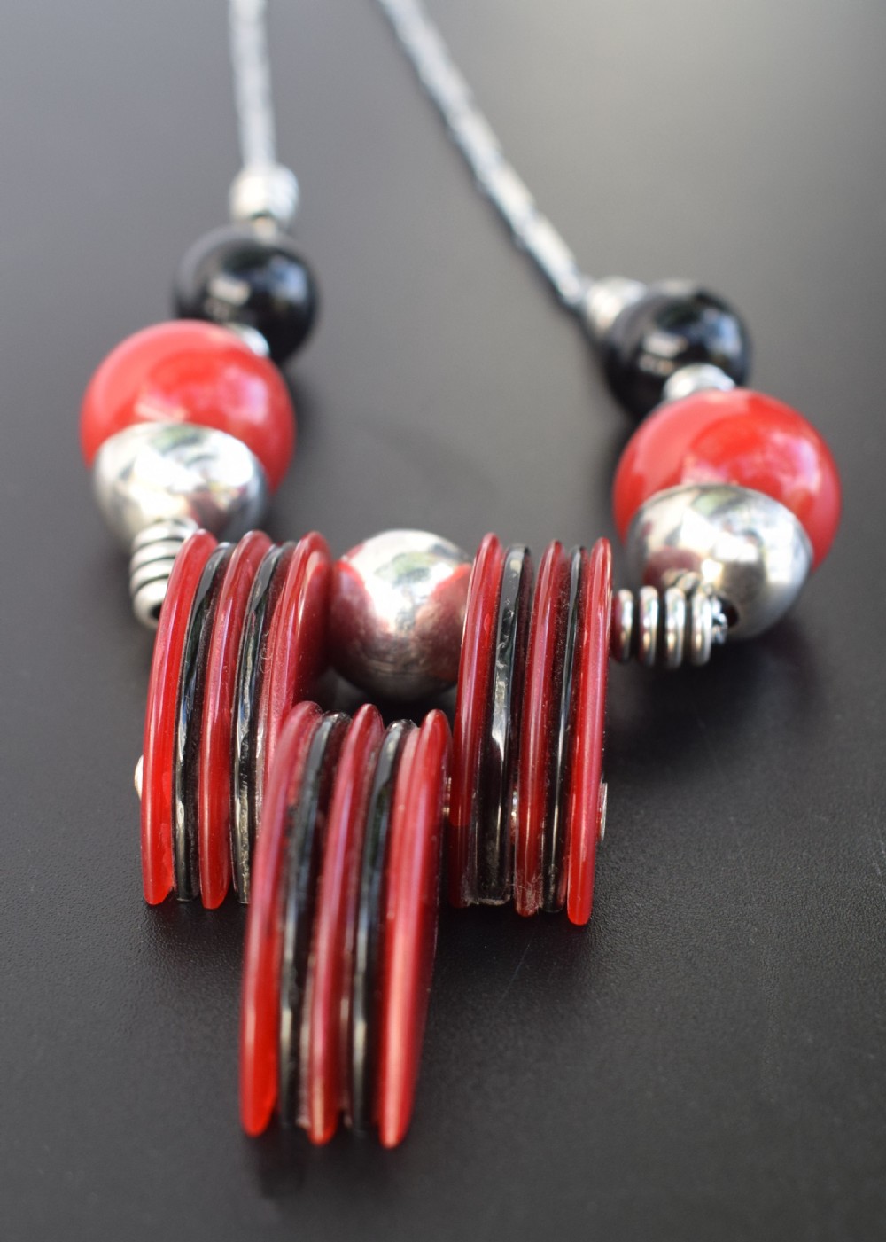An Art Deco modernist necklace featuring red and black spherical beads, coiled silver-toned metal spacers, and a geometric central pendant composed of stacked red and black discs on a metal link chain. View VI