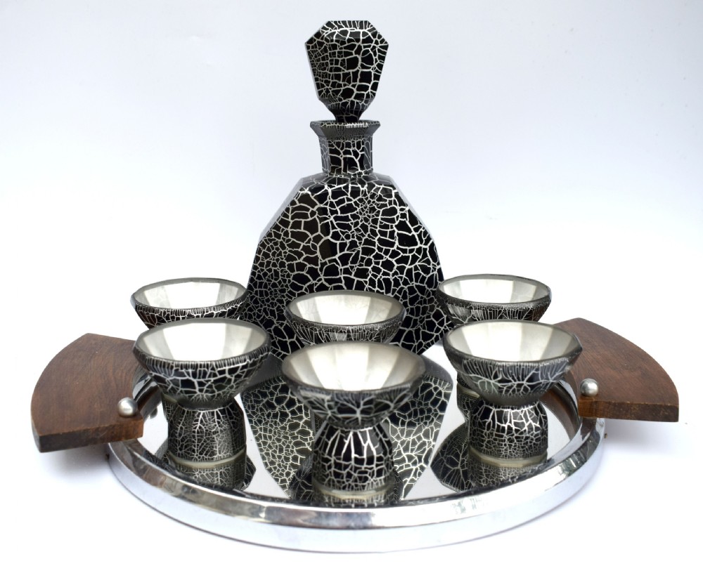 An Art Deco black and silver crackle-patterned glass decanter with six matching small cups arranged on a circular mirrored tray with wood handles.