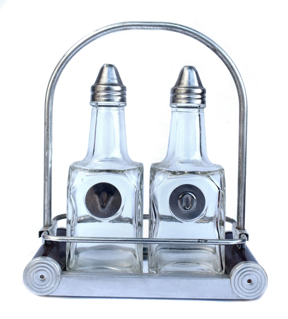 A pair of clear glass oil and vinegar bottles with silver metal tops marked V and O, housed in an Art Deco chrome carrying caddy.