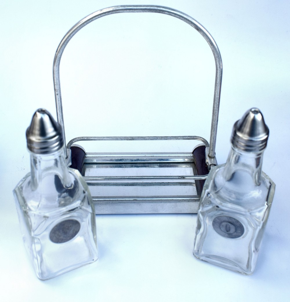 A pair of clear glass oil and vinegar bottles with silver metal tops marked V and O, housed in an Art Deco chrome carrying caddy. View V