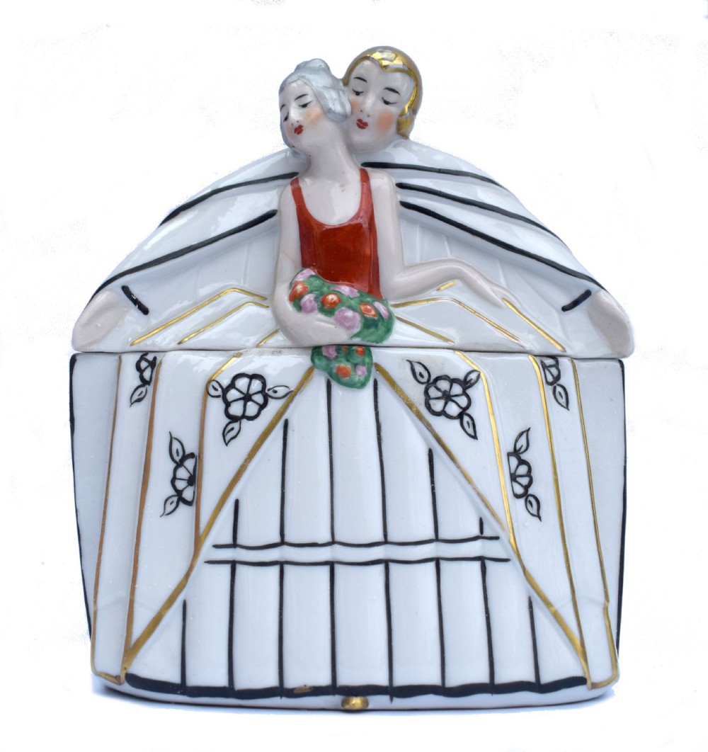 An Art Deco porcelain powder box featuring a lid sculpted in the form of an embracing couple in 1930s evening dress with black, red, and gold accents.
