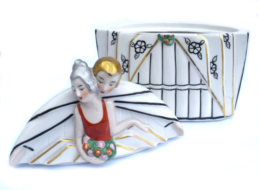 An Art Deco porcelain powder box featuring a lid sculpted in the form of an embracing couple in 1930s evening dress with black, red, and gold accents. View V