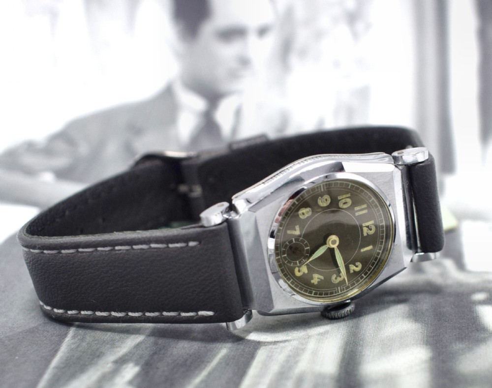 An Art Deco gents manual wristwatch featuring a metallic geometric case, a dark dial with Arabic numerals, and a dark brown leather strap with contrast stitching.