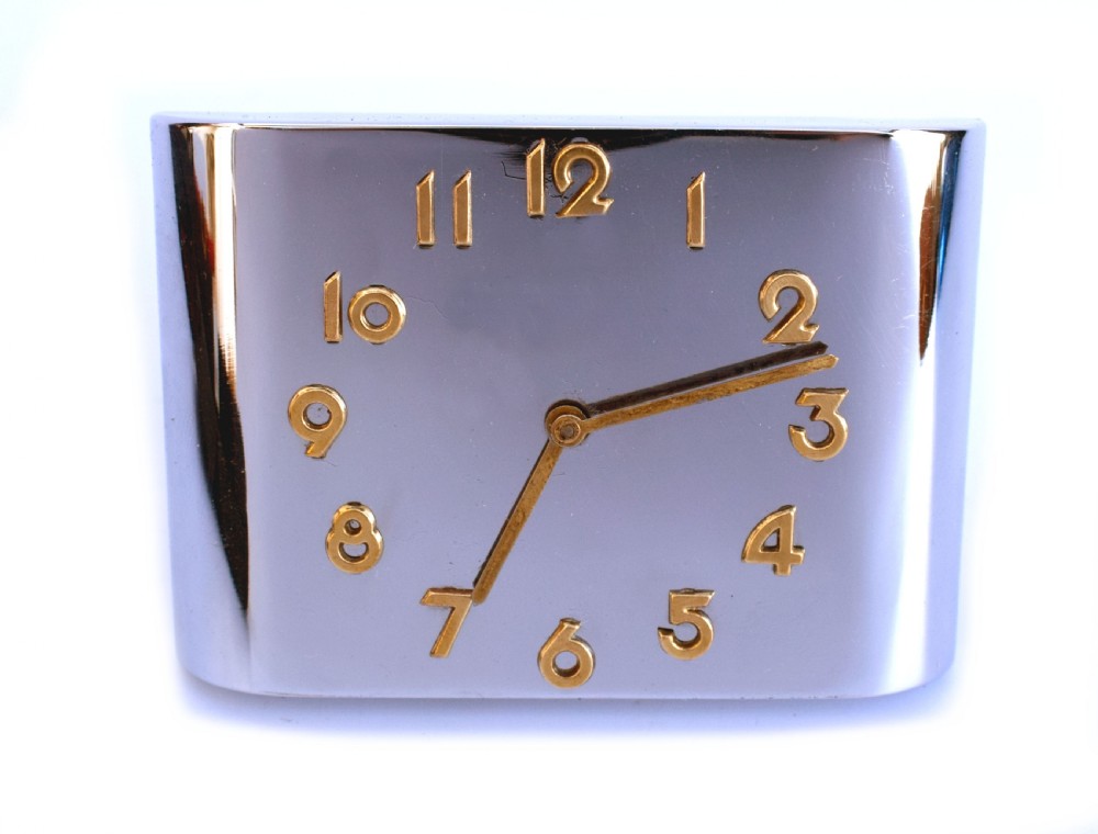 A curved rectangular chrome Art Deco travel clock featuring raised gold-coloured Arabic numerals and matching gold hands. View II