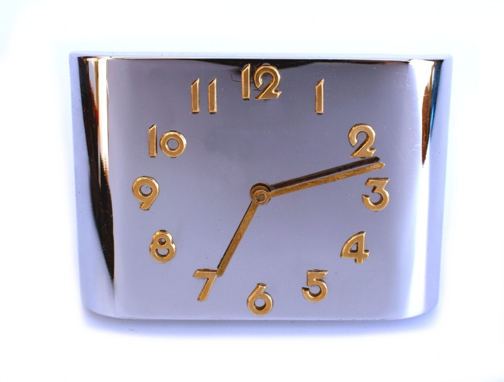 A curved rectangular chrome Art Deco travel clock featuring raised gold-coloured Arabic numerals and matching gold hands.