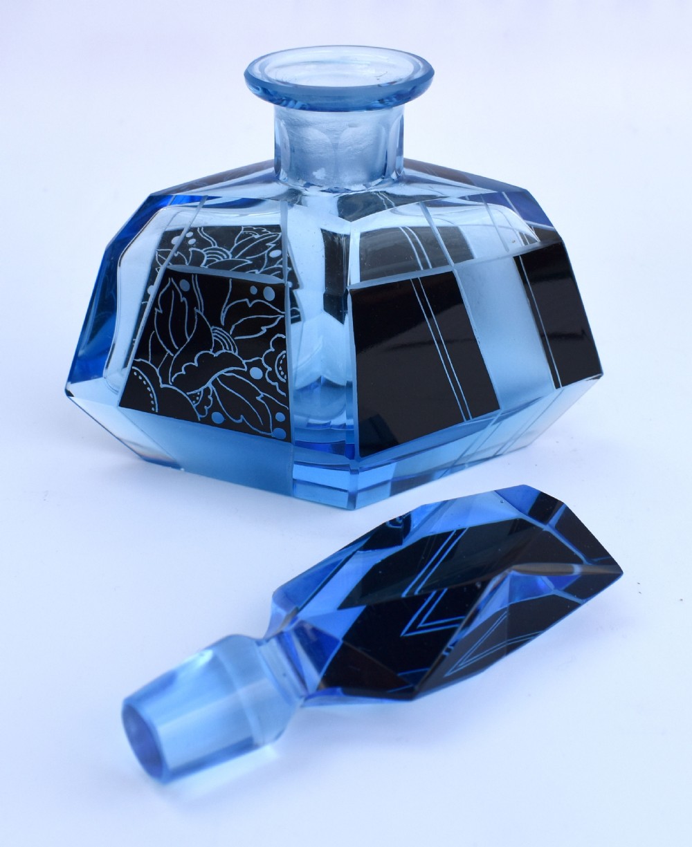 A blue Art Deco glass perfume bottle featuring geometric cut facets, black enamelled panels with etched floral designs, and a tall angular stopper. View VIII